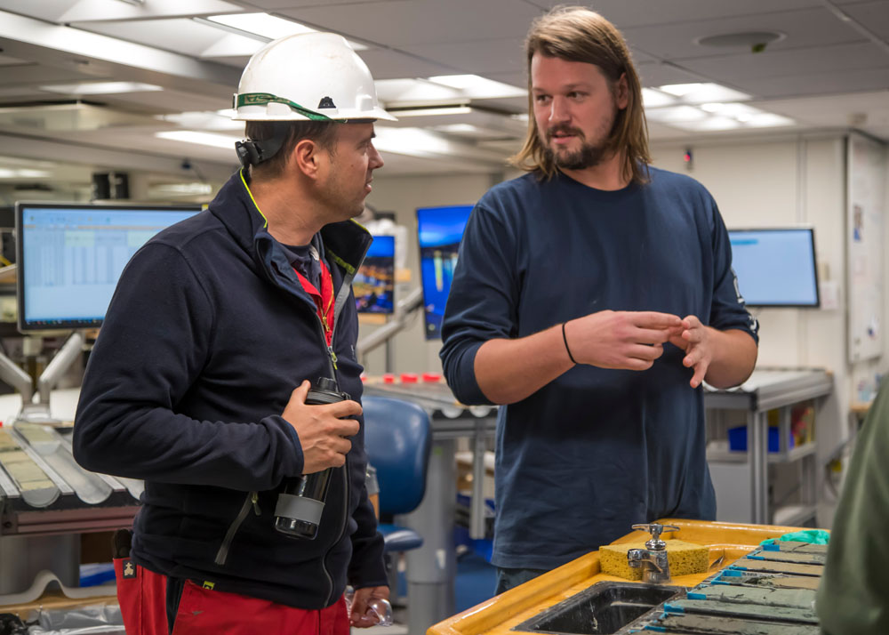 Siem Driller visits the core laboratory to view the split section halves he drilled that day. Dustin Harper (Sedimentologist, University of Kansas, USA) explains their features and geological background. (Credit: Sandra Herrmann, IODP JRSO) [Photo ID: exp396_131]